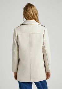 Beige wool blend coat with a structured design, large notched collar, and stitched accents. Features an identifiable label at the back.