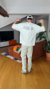 Person i hvit caps og overdimensjonert hvit skjorte som peker med tomlene mot logoen på baksiden av skjorten mens de står i en stue.