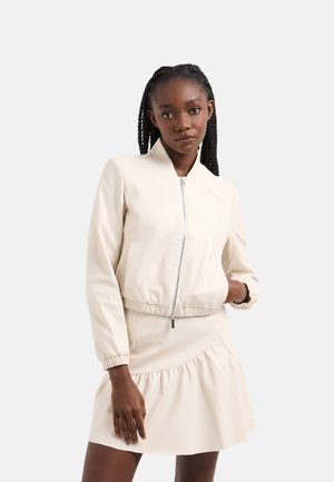 Giacca bomber cropped crema con zip frontale, vestibilità slim e polsini elasticizzati. Abbinata a un vestito con orlo a volant coordinato.