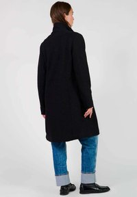 Manteau en laine noir avec un col haut, présentant des détails de couture, porté sur un jean bleu avec des manches retroussées et des mocassins noirs. Vue de dos affichée.