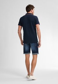 Polo navy con acentos rojos, combinado con pantalones cortos de mezclilla enrollados. La tela parece suave; el modelo lleva zapatos de color claro.