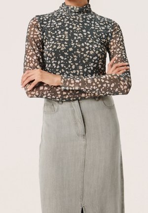 Woman wearing a high-neck, long-sleeve sheer black top with beige floral patterns and a high-waisted light gray skirt, arms crossed.