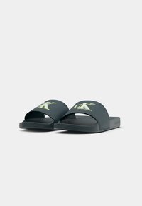 Sandales à glisser gris foncé avec un design à bout ouvert, présentant un détail de logo vert sur la sangle, fabriquées en matériau synthétique lisse.