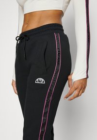 Pantalones de chándal negros con cintura ajustable, bolsillos laterales y franjas laterales en rosa contrastante que presentan texto de la marca, junto a un parche con el logo.
