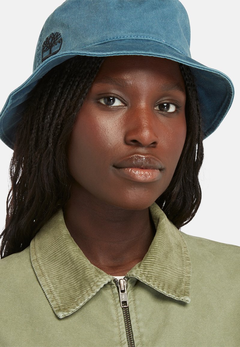 Cappello a secchi blu in suede con logo ricamato; indossato con una giacca in corduroy verde con chiusura con zip e colletto a costine.