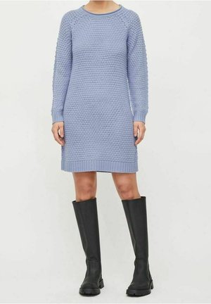 Kobieta ubrana w teksturowaną, jasnoniebieską sweterkową sukienkę w połączeniu z czarnymi kozakami do kolan, stojąca na tle jednolitego tła.
