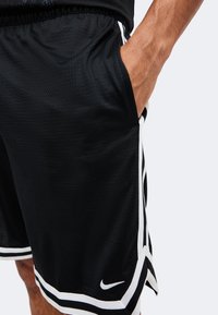 Gros plan d'une personne portant un short de sport Nike noir avec un liseré blanc, main reposant dans la poche avant.