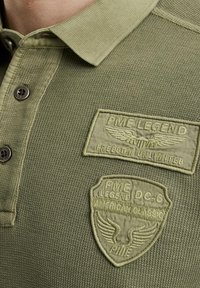 Olivgrünes Polohemd aus strukturiertem Stoff, mit Knopfverschluss und zwei gestickten Patches mit dem Markennamen und einem Flügel-Design.