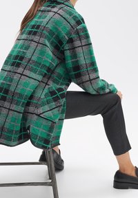 Person, der eine übergroße grün-grau karierte Jacke, schwarze Hosen und schwarze Slipper trägt, sitzt auf einem Metallhocker vor einem weißen Hintergrund.