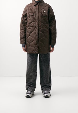 Persoon draagt een lange bruine doorgestikte jas met klepzakken, brede grijze spijkerbroek en zilveren sneakers tegen een effen lichte achtergrond.