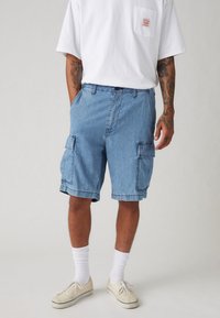 Denim cargo šortsevi u svijetloplavoj boji s dva velika bočna džepa, regularnog kroja i s prevrnutim rubom, uparen s bijelim majicom i cipelama.