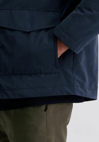 Mannenhand in de zak van een donkerblauwe jack met een klepzak, gekleed in olijfgroene broek op een grijze achtergrond.