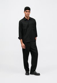 Svart skjorte med knapper, lomme og oppbrettede ermer, kombinert med løstsittende svarte bukser og svarte sko. Glatt tekstur.