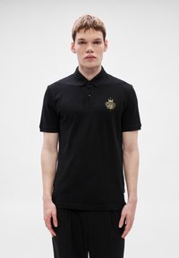 Melnas kokvilnas polo krekls ar divu pogu aizdari, īsām piedurknēm un zeltu izšūtu logotipu kreisajā krūtī.