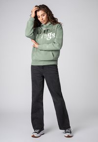 Vrouw in een saliegroene hoodie met de tekst "simple things", donkere wijde jeans en zwart-witte sneakers, poseert met haar hand op haar hoofd.