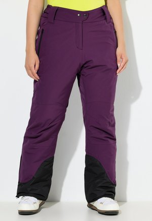 Personne portant un pantalon de plein air violet avec des poignets renforcés noirs et des bottes de neige blanches, se tenant devant un fond blanc.