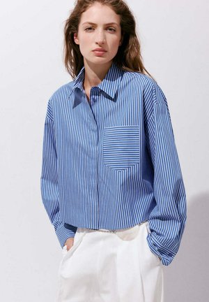 Femme portant une chemise à rayures verticales bleu et blanc avec une poche poitrine et un pantalon blanc, mains dans les poches, sur un fond uni.