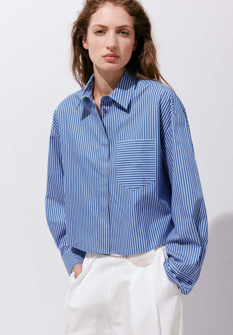 Femme portant une chemise à rayures verticales bleu et blanc avec une poche poitrine et un pantalon blanc, mains dans les poches, sur un fond uni.