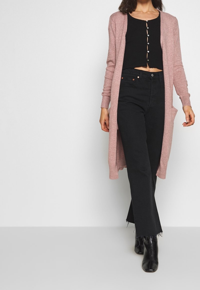 Lange, lichtroze open cardigan met ribbelstructuur, gedragen over een zwart crop top en zwarte hoge taille uitlopende jeans, gecombineerd met zwarte laarzen.