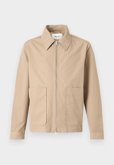 Veste beige à fermeture éclair avec col pointu, manches longues et deux poches avant, présentée sur un fond uni.