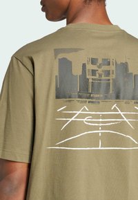 T-shirt di cotone verde oliva con un design grafico di uno skyline urbano e linee di un campo da basket in bianco e nero sul retro.