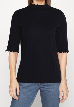 Femme portant un haut noir côtelé ajusté à manches courtes avec des bords festonnés et un jean bleu, debout devant un fond uni.