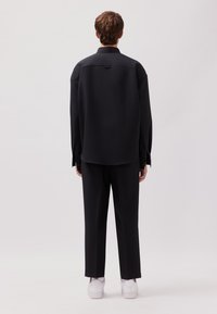 Schwarzes, oversized Hemd mit Kragen, Rückenbelüftung und langen Ärmeln, kombiniert mit schwarzen Hosen und weißen Sneakers. Glatter Stoff, minimalistisches Design.