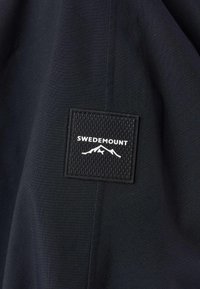 Tissu noir à surface texturée avec un patch carré portant l'inscription "SWEDEMOUNT" et un graphisme de montagne en blanc.