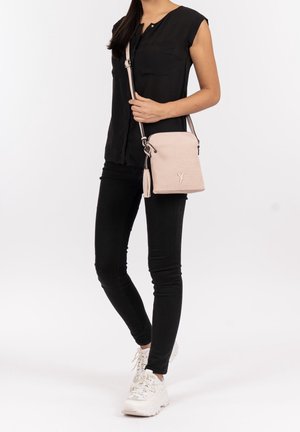 Donna in blusa senza maniche nera e pantaloni, che indossa sneakers bianche e porta una borsa a tracolla beige con dettaglio a frange.