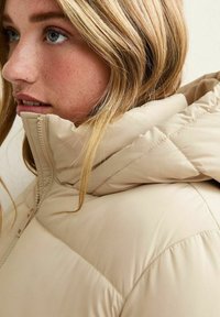 Close-up van een vrouw met blond haar die een beige gewatteerde jas draagt met een hoge ritssluiting en een capuchon.