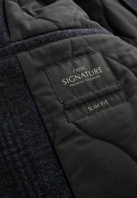 Must mustrilise kangas, millel on tekstuuriga villane välimine kiht. Peal on silt, millel on kuldsete tähtedega trükitud "next SIGNATURE PREMIUM TAILORING" ja "SLIM FIT".