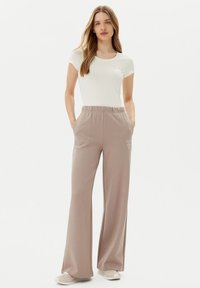 Pantaloni ampi color beige chiaro con vita elastica e tasche laterali, abbinati a una maglietta bianca a maniche corte semplice con dettaglio del logo.