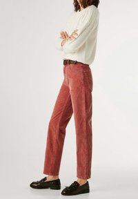 Personne debout, les bras croisés, portant un pull blanc, un jean droit rouge, une ceinture noire et des mocassins noirs sur fond uni.