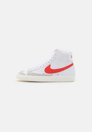 Witte hoge sneaker met rode Nike-swoosh, witte veters, grijze suède neuskap en off-white zool op een effen achtergrond.