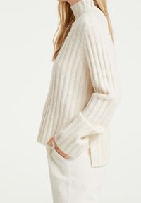 Pull côtelé de couleur crème avec un col montant, fabriqué en matériau doux. Présente une coupe décontractée et des poignets légèrement évasés.