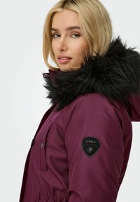 Parka bordeaux con cappuccio in pelliccia sintetica nera, chiusura con zip e bottoni, due tasche frontali e una patch rotonda con logo sulla manica sinistra.
