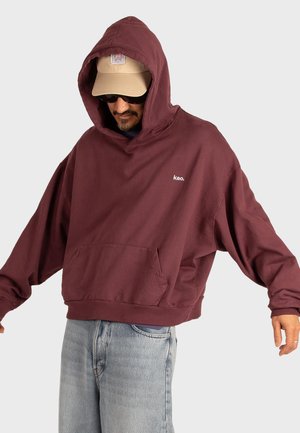 Bordeauxfarbener Oversized-Hoodie mit einer vorderen Tasche und einer Kapuze, die ein kleines weißes Logo zeigt. Kombiniert mit hellblauen Jeans.