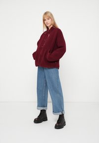 Naine, kes kannab burgundia värvi fliisdžeket, laiaid siniseid teksaseid ja musti šnörkidega platvormikingi, seisab valge tausta ees.