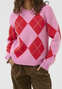 Personne portant un pull rose avec de grands losanges rouges argyle et un pantalon en velours côtelé marron, les mains dans les poches sur un fond neutre.
