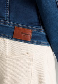 Džínová bunda s tmavě modrou látkou a hnědou koženou nálepkou s nápisem "Pepe Jeans London." Pod tím je krémová kapsa s texturovanou látkou.