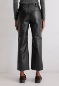 Schwarze Leder-Hose im weiten Schnitt mit glatter Textur, mit Gesäßtaschen und hohem Bund, getragen mit braunen Schuhen auf einer hellen Oberfläche.