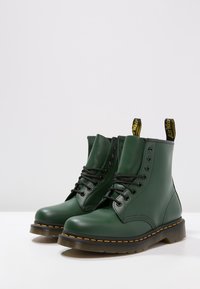 Zielone skórzane botki za kostkę z czarnymi sznurówkami, okrągłym czubkiem, przeszyciami, żółtą pętelką i kontrastowym żółtym przeszyciem na podeszwie.