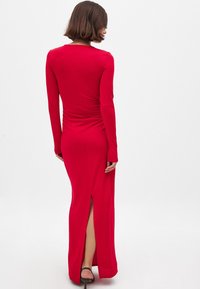 Red long-sleeve dress with a fitted design, ruched detailing at the back, and a thigh-high slit, made from smooth, stretchy fabric.