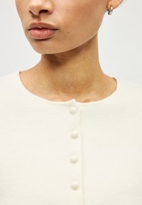 Krēmkrāsas kardigans ar apaļu izgriezumu un pieciem lieliem apaļiem pogām priekšpusē. Mīksts, teksturēts audums ar gludu apdari.