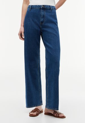 Person iført blå jeans med høj talje og lige ben samt brune flade sandaler med stropper, stående mod en ensfarvet hvid baggrund.
