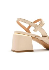 Sandalia de vestido beige con tacón cuadrado. Presenta correas entrelazadas y un hebilla, mostrando una textura de piel suave y costuras mínimas.