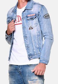 Ljusblå jeansjacka med en blekt tvätt, prydd med lappar på bröstet och ärmen, samt en klassisk knappuppläggning på framsidan.