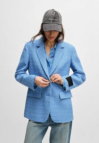 Hellblau strukturierter Blazer mit lockerer Passform, eingekerbten Revers, Fronttaschen und einem schwarzen Akzent am Ärmel. Kombiniert mit einem blauen Hemd und einer Kappe.