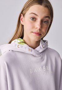 Lys lilla hættetrøje lavet af blødt stof, med indvendig hætte med blomstermønster og prægede hvide logo. Guld hardware-accenter.