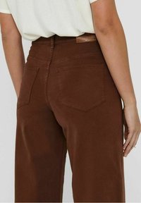 Persoon in hooggerande bruine broek met achterzakken en een ingestopt wit shirt, weergegeven vanaf de taille naar beneden van achteren.